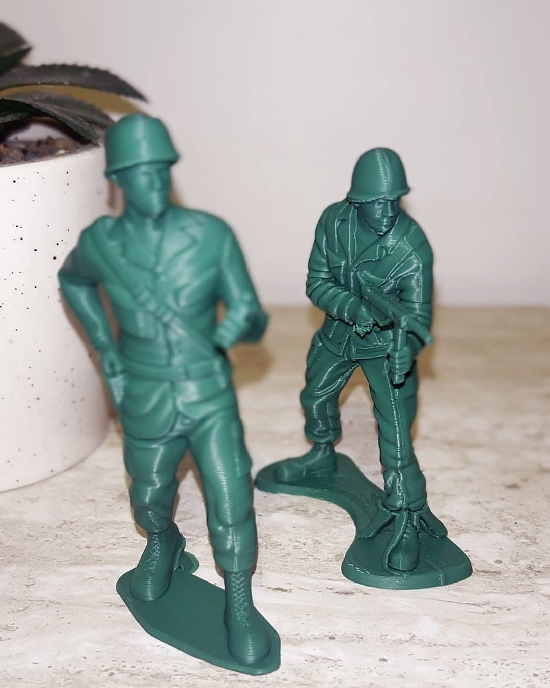 Two green PLA army men with their supports removed, on a marble shelf beside a potted plant