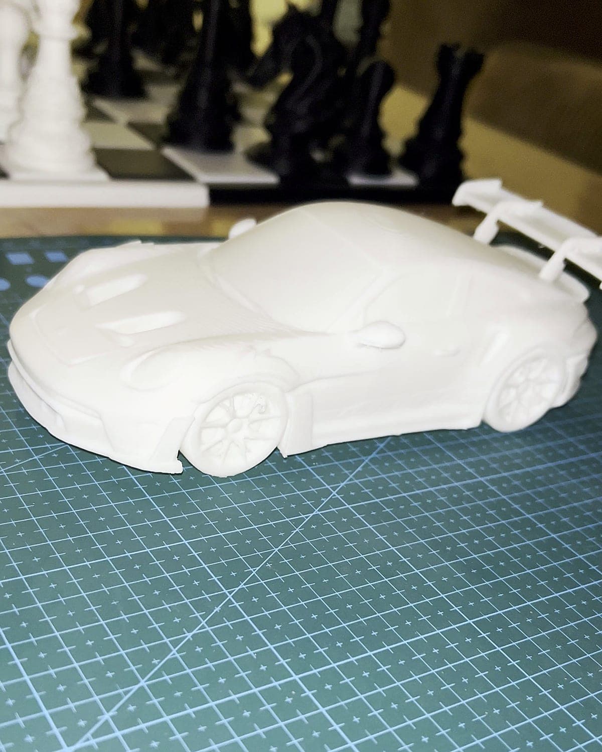 The whole white PLA sports car model, rear wing and wheels included, on a green cutting mat with a chess set behind