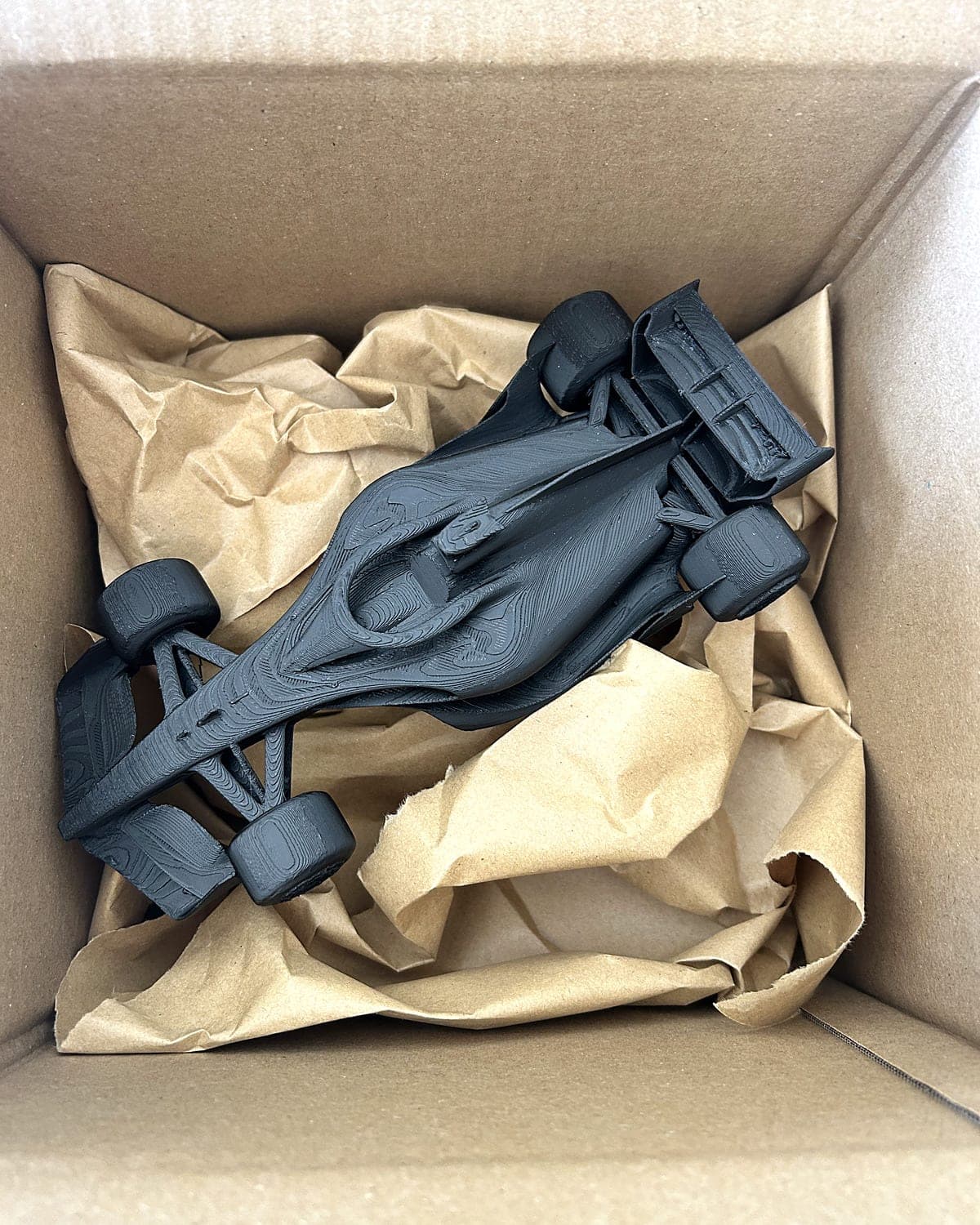 Black PLA race car model lying in crumpled brown paper inside a cardboard box, packed to courier