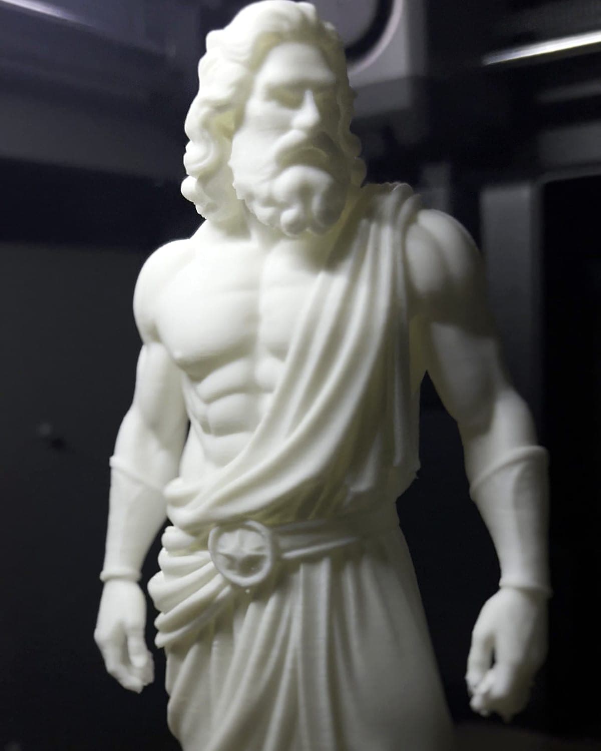 Low-angle view of the white PLA Zeus figurine, showing the carved beard, muscles and draped robe