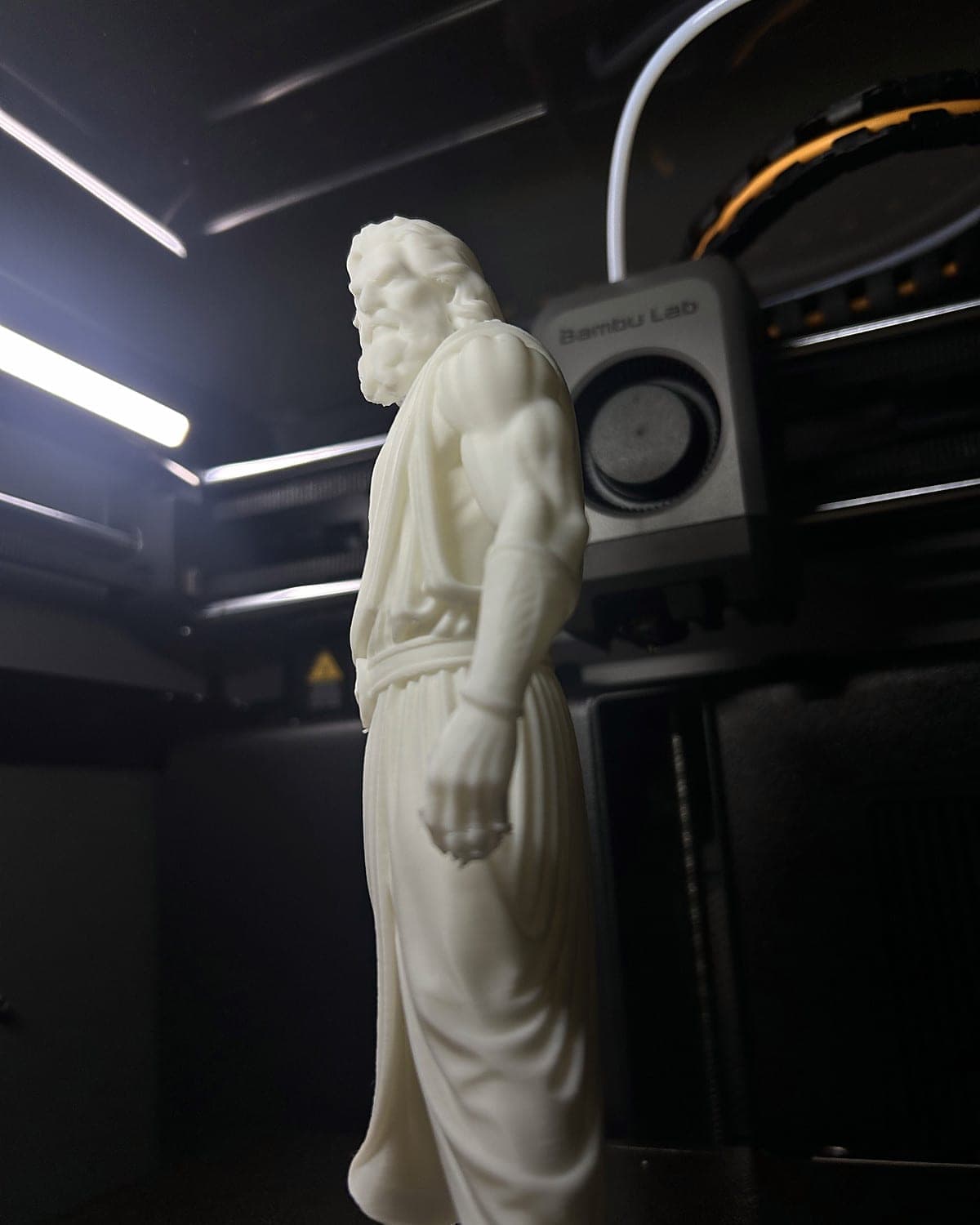 The white Zeus figurine standing inside a Bambu Lab 3D printer under its LED light strip