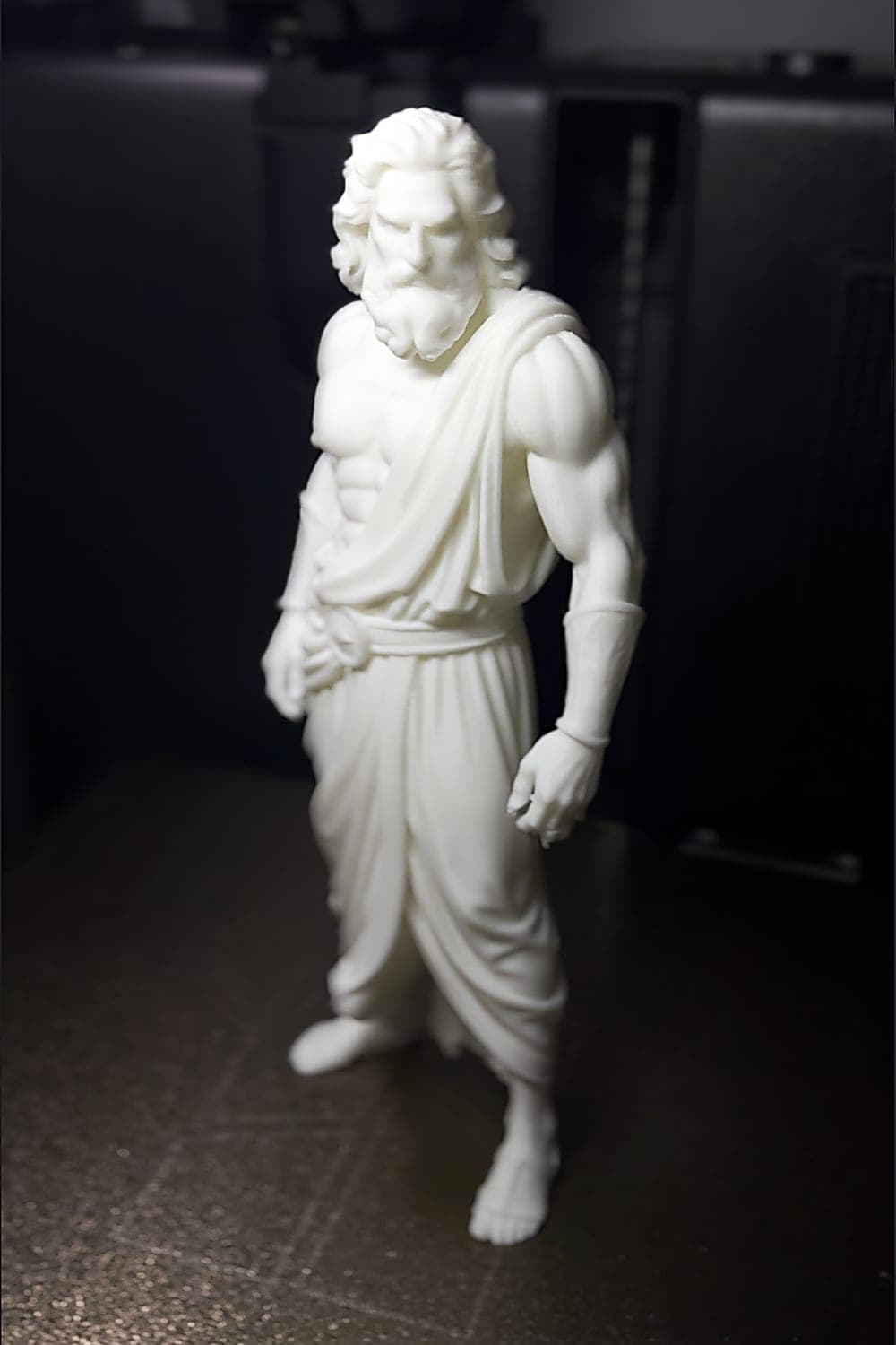 The Zeus figurine 3D-printed in white PLA, standing full length on a dark print bed