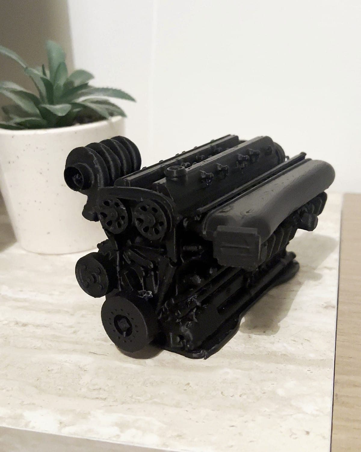Black 3D-printed model of a car engine on a marble shelf beside a potted succulent