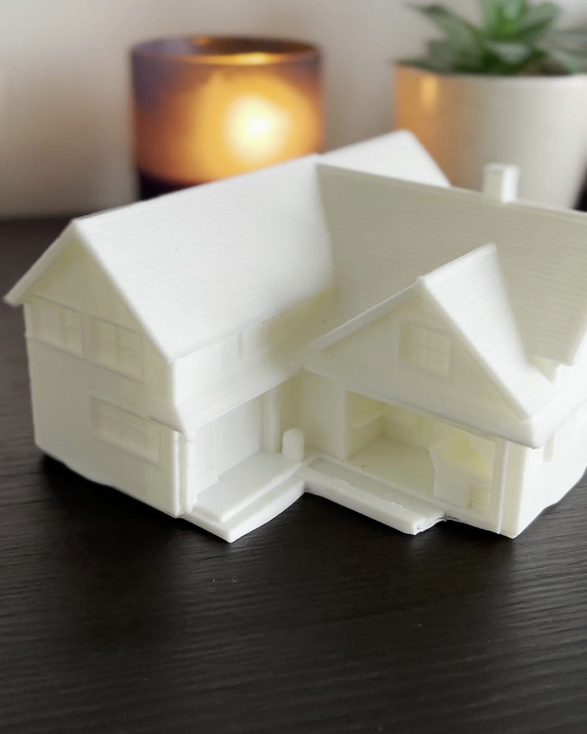 White 3D-printed model of a two-storey house on a dark wooden table, a lit candle and a pot plant behind it